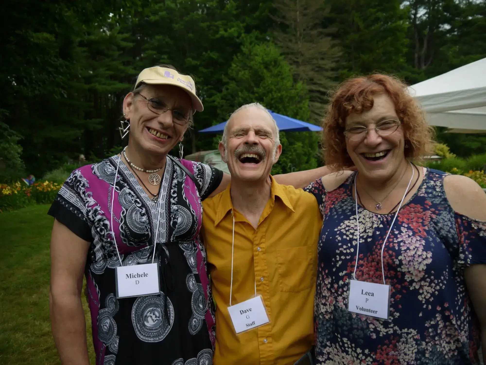 Two women in floral shirts and one man in a mustard collared shirt laugh with their arms around one another.