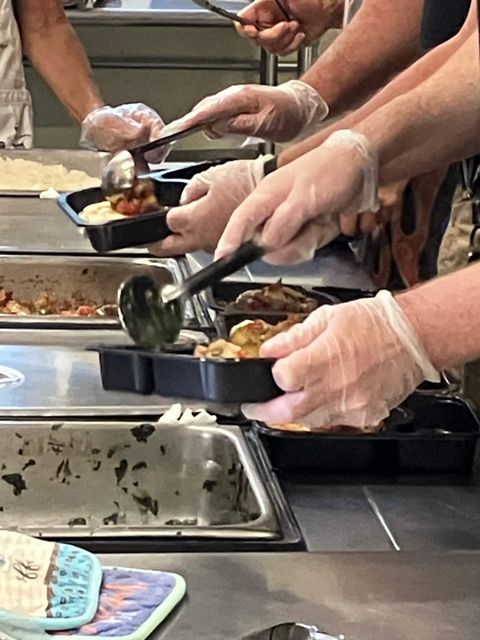 Many gloved hands dishing food into Meals on Wheels trays.