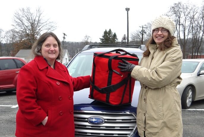 Jane Severance, Nutrition Program Director (left), standing in front of a car, holding an insulated bag for home-delivered meals, with Lynne Feldman, Director of Community Services