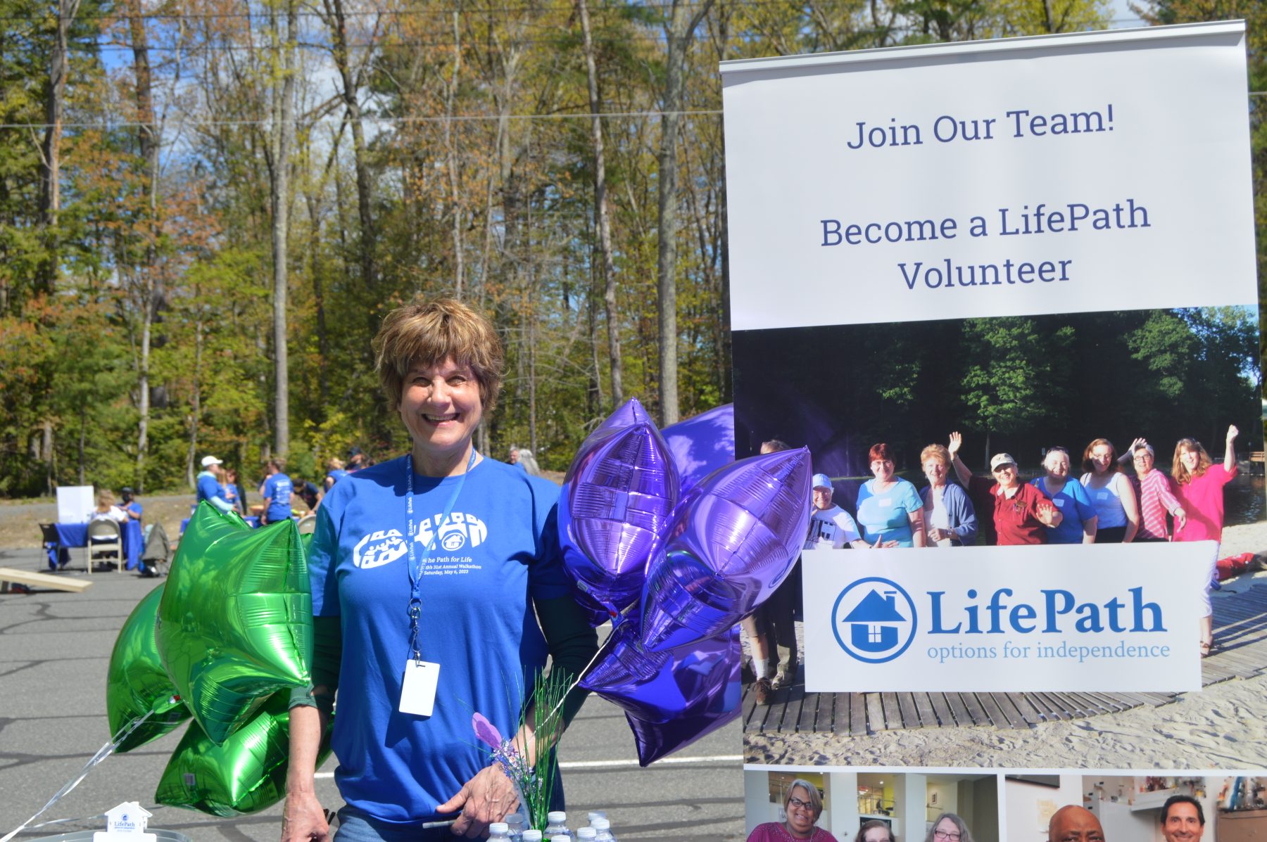 A LifePath staff member stands with balloons and a sign saying "Join Our Team! Become a LifePath Volunteer."