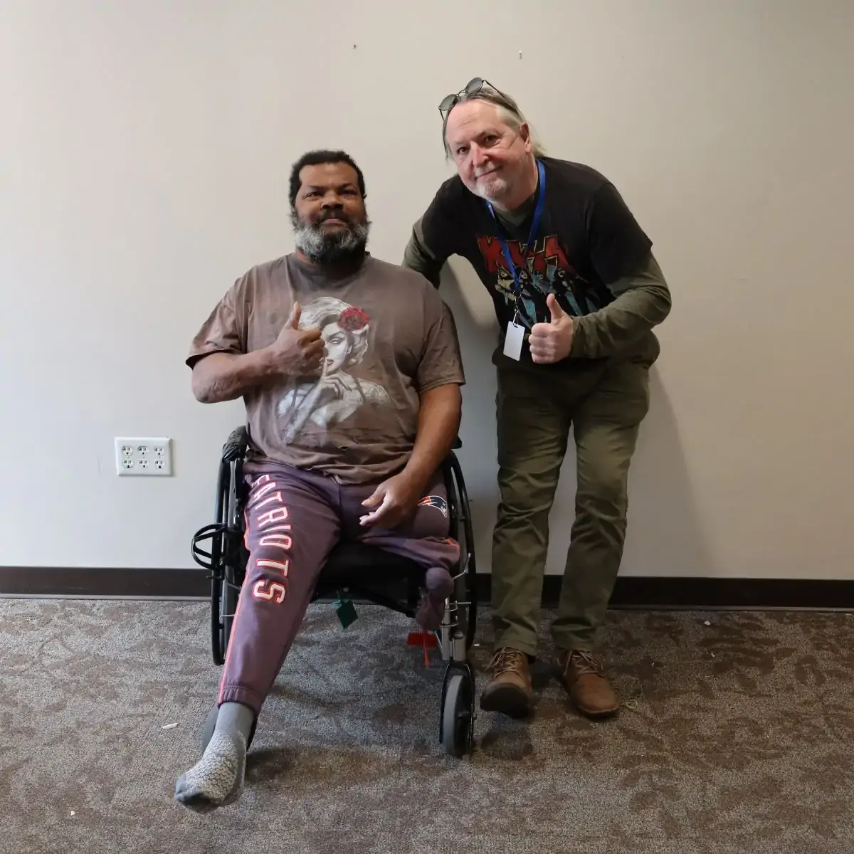 Damon in a wheelchair and Peter-Michael leaning by chair, both making thumbs up sign