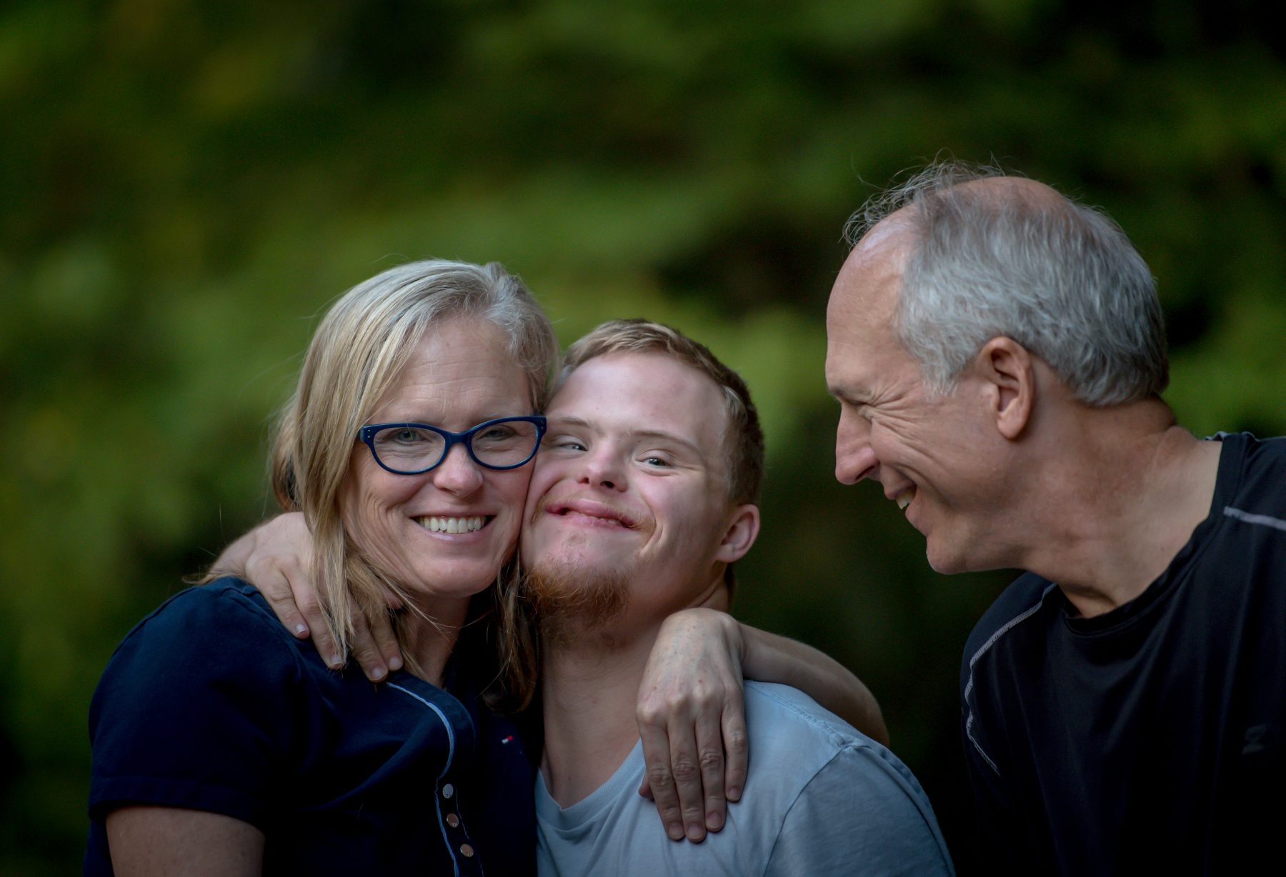 Mother and father hugging young man with Down Syndrome outside.