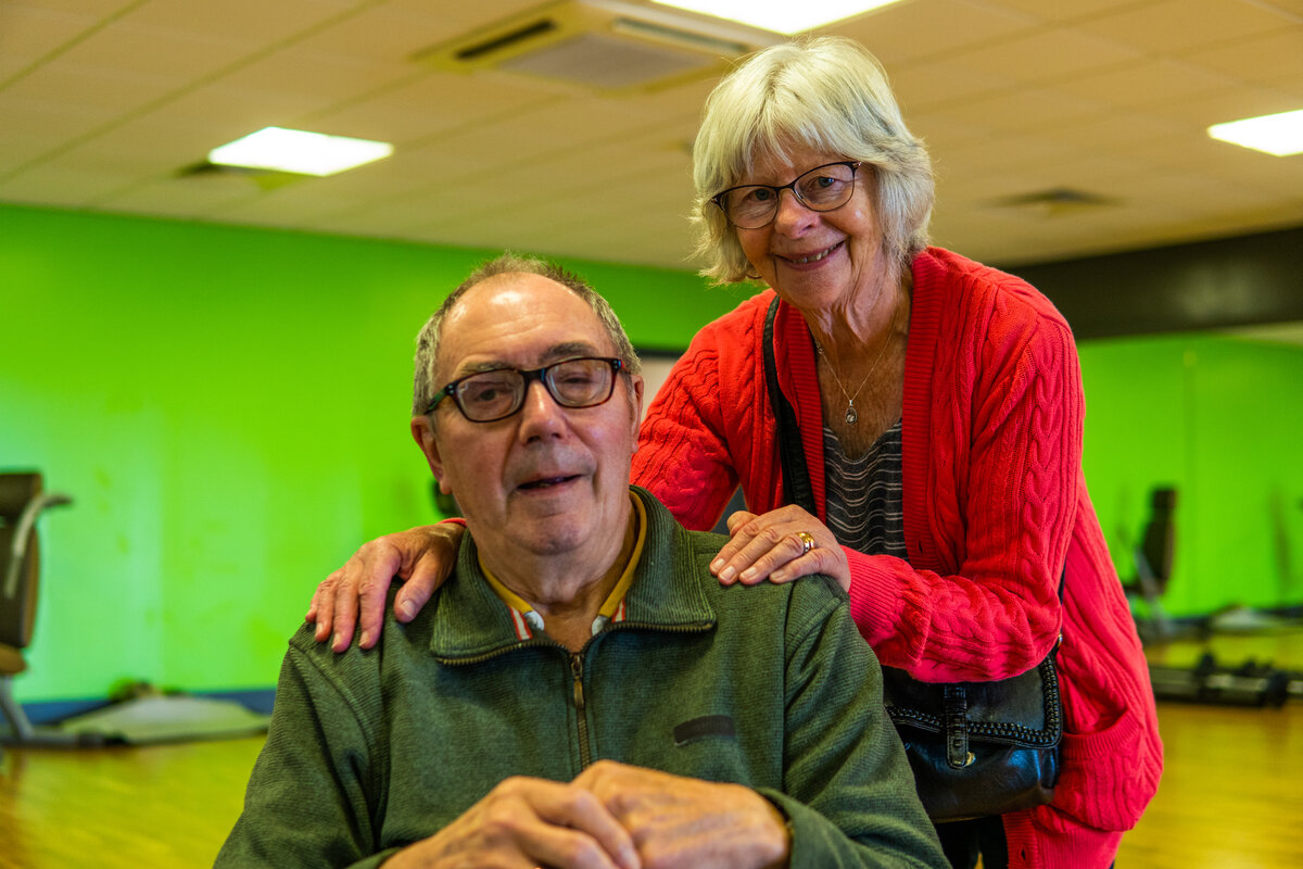 older woman smiling with hands on shoulders of sitting older man