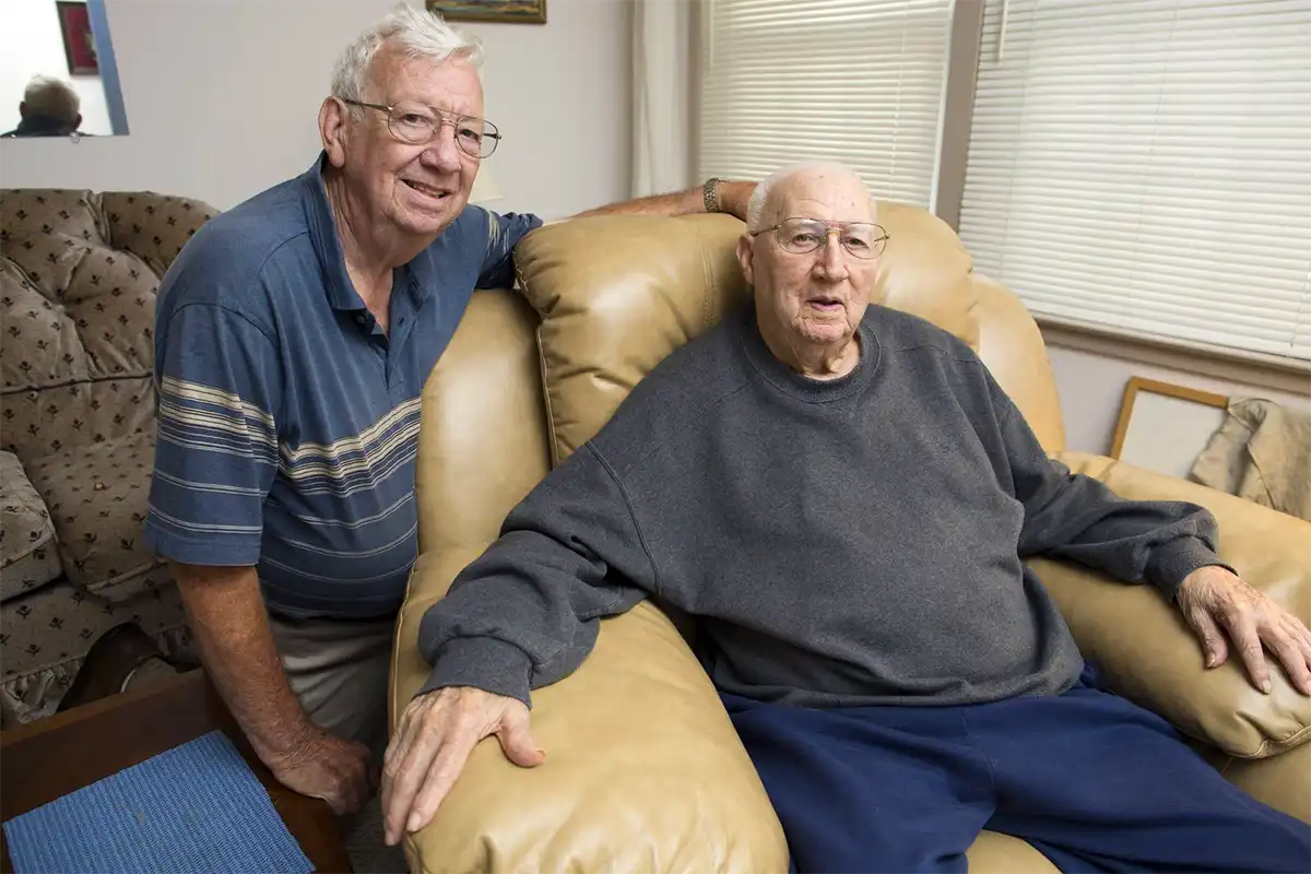 An older adult sits in a comfortable chair while another man puts his arm around the back