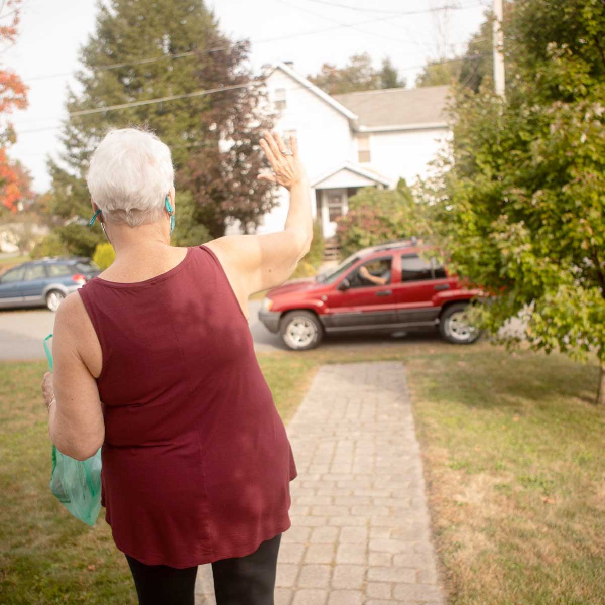 A woman waves from her doorstep to her Meals on Wheels driver