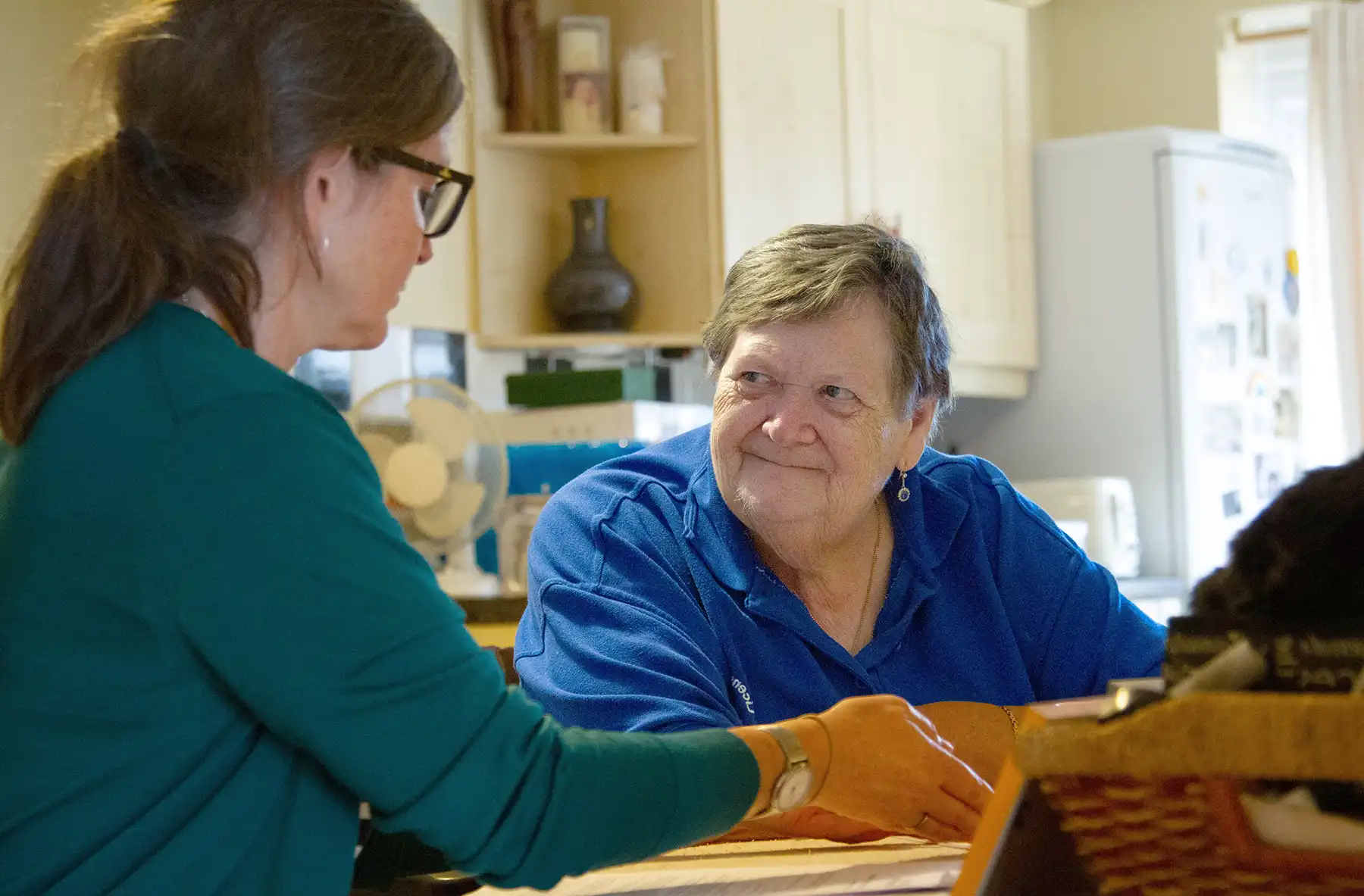 An older adult woman sits at her kitchen table with a younger woman